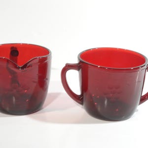 Puede incluir: Un juego de dos jarras de crema y azucarero de vidrio rojo vintage. La jarra de crema tiene un asa y el azucarero tiene dos asas.