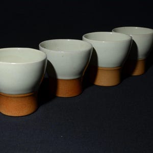 May include: Four white ceramic cups with a contrasting orange-brown base are arranged in a row. The cups have a simple, elegant design, with a slightly flared rim and a smooth, matte finish. The background is a dark, solid color.