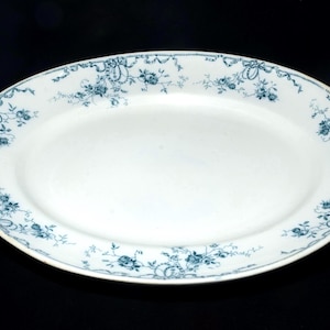 May include: An oval-shaped white ceramic serving platter featuring a decorative border pattern of dark teal floral motifs, delicate ribbons, and swags that encircle the entire rim of the dish, leaving the centre of the platter plain and white.
