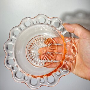 Pode incluir: Uma tigela de vidro rosa com uma borda festonada e um design vazado. A tigela tem um centro claro e facetado.