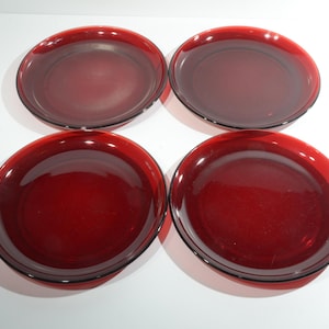 Puede incluir: Cuatro platos redondos de vidrio rojo rubí. Los platos tienen un borde ligeramente elevado y una superficie lisa y brillante. Los platos están dispuestos en forma cuadrada, perfectos para servir aperitivos o postres.