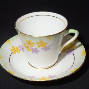 May include: A white teacup and saucer with a floral pattern. The cup and saucer have a gold trim and feature yellow, purple, and green flowers.