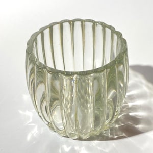 Op de afbeelding: Transparante glazen votiefkaarsenhouder met verticaal geribbeld ontwerp. De kleine, ronde houder heeft een geschulpte rand. Het transparante glas laat licht door en werpt een schaduw.