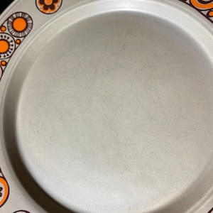 May include: A beige ceramic plate with a brown and orange geometric pattern around the rim. The pattern features circles, swirls, and flowers.