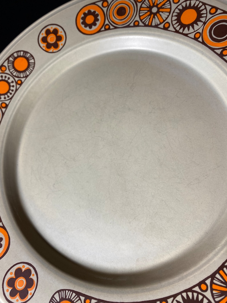 May include: A beige ceramic plate with a brown and orange geometric pattern around the rim. The pattern features circles, swirls, and flowers.