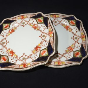 May include: Two square-shaped ceramic plates with a scalloped edge. The plates have a white background with a floral pattern in shades of red, orange, yellow, green, and blue. The center of each plate is a white circle.