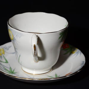 May include: White teacup and saucer set with a floral design. The cup and saucer have a gold trim and a delicate handle. The floral pattern includes yellow, red, and green accents. The teacup is sitting on a matching saucer.