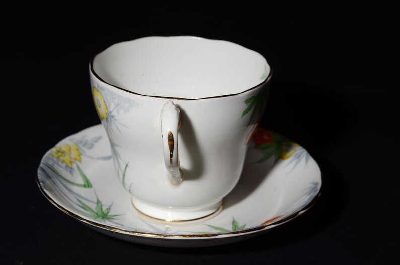 May include: White teacup and saucer set with a floral design. The cup and saucer have a gold trim and a delicate handle. The floral pattern includes yellow, red, and green accents. The teacup is sitting on a matching saucer.