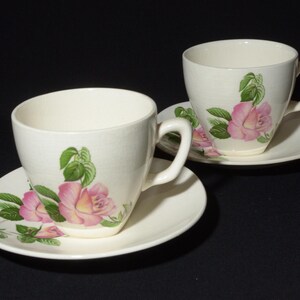 May include: Two white teacups with pink rose floral patterns and matching saucers. The cups have a delicate floral design with pink roses and green leaves.
