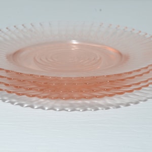 Puede incluir: Una pila de cuatro platos de vidrio rosa. Cada plato tiene un dise&ntilde;o en espiral en el centro y un borde estriado y festoneado. Los platos est&aacute;n apilados uno encima del otro, con el plato superior enfocado.