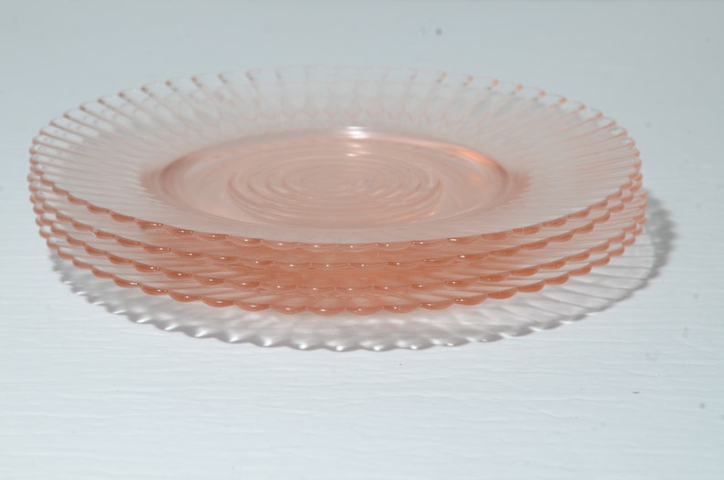 Puede incluir: Una pila de cuatro platos de vidrio rosa. Cada plato tiene un dise&ntilde;o en espiral en el centro y un borde estriado y festoneado. Los platos est&aacute;n apilados uno encima del otro, con el plato superior enfocado.