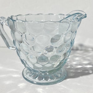 Puede incluir: Una jarra de crema de vidrio transparente con una superficie texturizada y con burbujas. La jarra tiene un asa y una base pequeña y redonda.