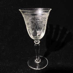 May include: A clear, etched glass goblet with a long stem and a wide bowl. The glass features a detailed pattern around the rim and on the bowl. The goblet is displayed against a dark background, highlighting its delicate design.