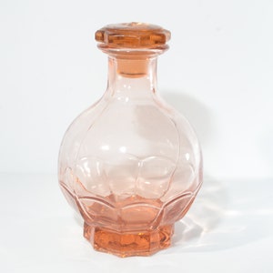 May include: A clear glass bottle with a pink hue and a textured, faceted design. The bottle has a matching pink glass stopper.