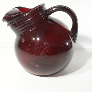 Puede incluir: Una jarra de vidrio de color rojo oscuro con cuerpo redondo y asa. La jarra tiene un diseño texturizado alrededor de la parte superior y un diseño de uva grabado en el lateral.