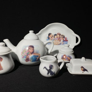 Puede incluir: Un juego de té de porcelana blanca con tetera, jarra de crema, azucarero, mantequillera y bandeja. La tetera tiene un diseño floral con una niña sosteniendo un ramo de flores. La jarra de crema tiene un diseño de gato negro. El azucarero tiene un diseño de dachshund. La mantequillera tiene un diseño de pájaro. La bandeja tiene un diseño de niños comiendo una tarta.