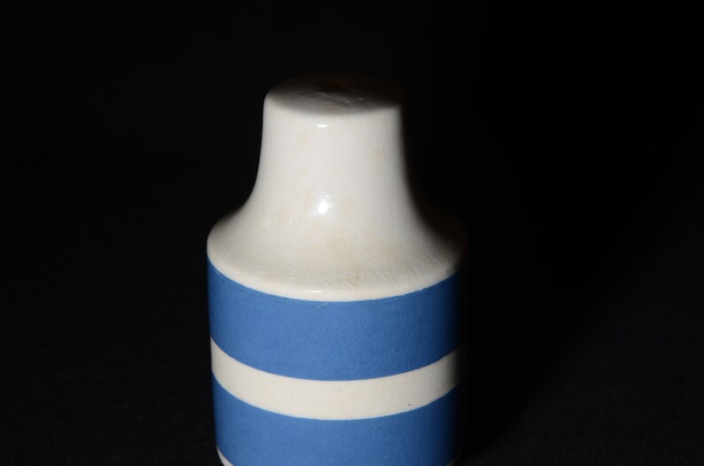 May include: A white ceramic salt shaker with blue stripes. The shaker has a round body and a small opening at the top.