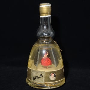 May include: A clear glass Bols bottle with a gold cap and label. Inside the bottle is a miniature figurine of a woman in a red dress. The label features the word "BOLS" and a portrait.
