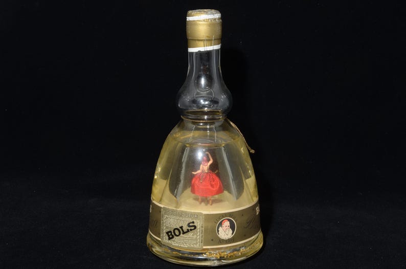 May include: A clear glass Bols bottle with a gold cap and label. Inside the bottle is a miniature figurine of a woman in a red dress. The label features the word "BOLS" and a portrait.