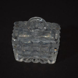 May include: A clear glass bottle with a square base and a textured design. The bottle has a round top with a small opening.