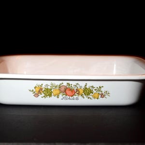 Puede incluir: Fuente rectangular blanca para hornear con bordes redondeados. El plato presenta un colorido diseño floral y de frutas con el texto "L'échalote" en la parte delantera. El plato está sobre un fondo negro.