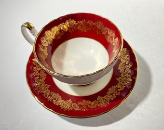 May include: A red teacup and saucer with gold trim and a floral pattern. The cup and saucer are both decorated with a delicate gold design.