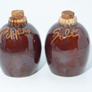 May include: Two brown ceramic salt and pepper shakers. The round shakers have cork stoppers and the words "Pepper" and "Salt" in a lighter brown script. The shakers have a glossy finish.