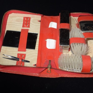 May include: A red zippered travel grooming kit containing a rectangular mirror, metal tweezers, two white combs, a black rectangular container, and two round brushes with white bristles, all secured within red elastic straps inside the case.