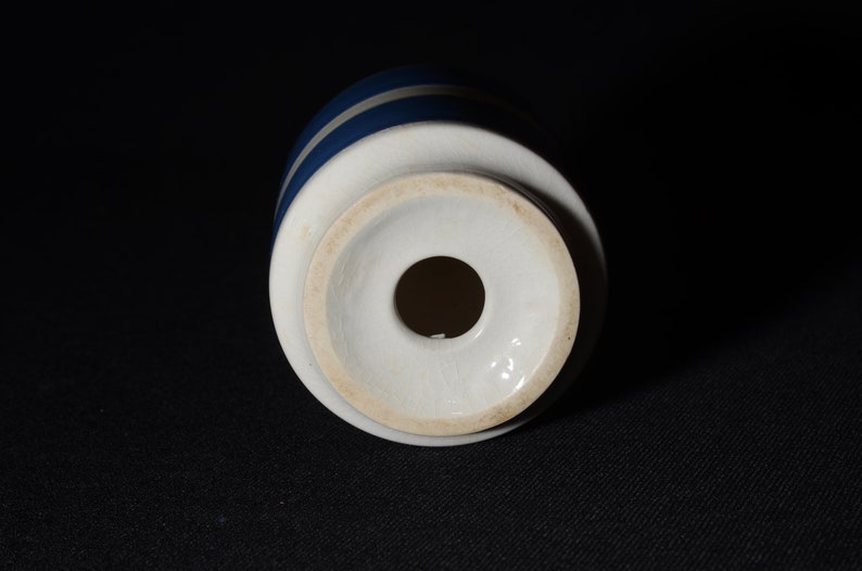 May include: White ceramic bottle stopper with a blue and white striped design. The stopper has a round hole in the center.