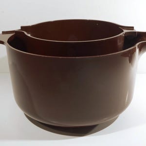 May include: Two nested, dark brown mixing bowls. The bowls are made of a glossy material and have handles on either side. The bowls are designed to nest inside each other for storage.