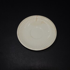 May include: A white ceramic saucer with a fluted design. The saucer is empty and has a slight yellow tint.