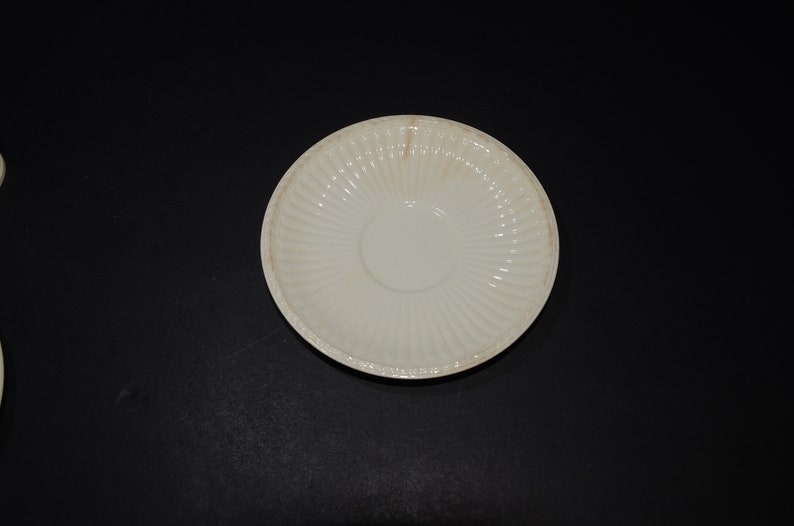 May include: A white ceramic saucer with a fluted design. The saucer is empty and has a slight yellow tint.