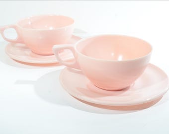 Puede incluir: Dos tazas de té y platillos rosas. Las tazas están hechas de un material liso y opaco y tienen una forma redondeada. Los platillos son ligeramente más grandes que las tazas y tienen un fondo plano.