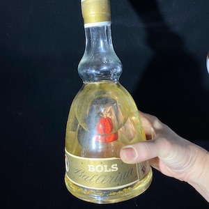 May include: A vintage Bols Ballerina bottle with a gold cap and label. The clear glass bottle contains a red ornament and a yellow liquid. The label reads "Bols Ballerina" in gold lettering.