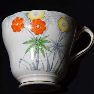 May include: White teacup with a floral design featuring orange and yellow flowers, green leaves, and gray accents. The cup has a gold rim and handle, and is set against a dark background. A decorative item for tea lovers.