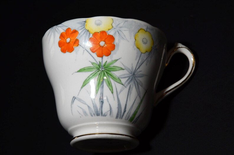 May include: White teacup with a floral design featuring orange and yellow flowers, green leaves, and gray accents. The cup has a gold rim and handle, and is set against a dark background. A decorative item for tea lovers.