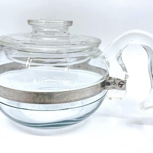 May include: A clear glass teapot with a matching glass lid and a curved glass handle. A metallic band encircles the centre of the round body, featuring the word STAINLESS embossed on the side. The design includes a wide, rounded base.