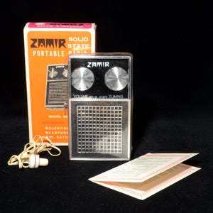 May include: A vintage Zamir solid state portable radio with a silver and black face, two circular tuning knobs, and a grid speaker pattern. The set includes an orange cardboard box, a white wired earphone, and a folded paper instruction manual.