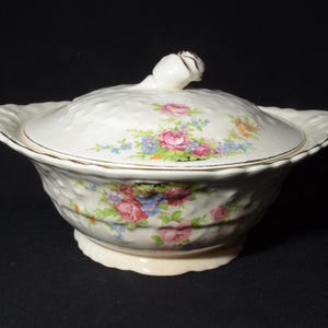 May include: A white ceramic serving bowl with a lid. The bowl and lid are decorated with a floral pattern of pink, red, and blue roses and other flowers. The lid has a rosebud finial.