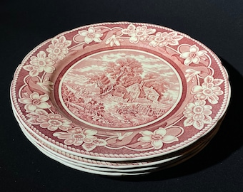 Pode incluir: Um conjunto de quatro pratos de jantar de cerâmica rosa e branca com uma borda floral e uma imagem central de uma casa de campo em uma paisagem.