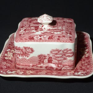 May include: A white ceramic butter dish with a red and white floral pattern. The dish has a square base and a lid with a knob on top. The dish is decorated with a floral pattern and a scene of a bridge and trees.