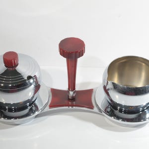 Puede incluir: Un juego de azucarero y cremera de cromo con detalles en plástico rojo. El juego incluye un asa central con un pomo rojo y dos cuencos redondos con tapas.