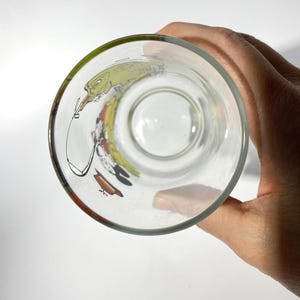 K&ouml;nnte beinhalten: Klares Glas mit einem Cartoon-Charakter-Design. Das Glas hat einen gr&uuml;nen Rand und eine farbenfrohe Illustration auf der Innenseite. Das Glas wird von einer Hand gehalten.