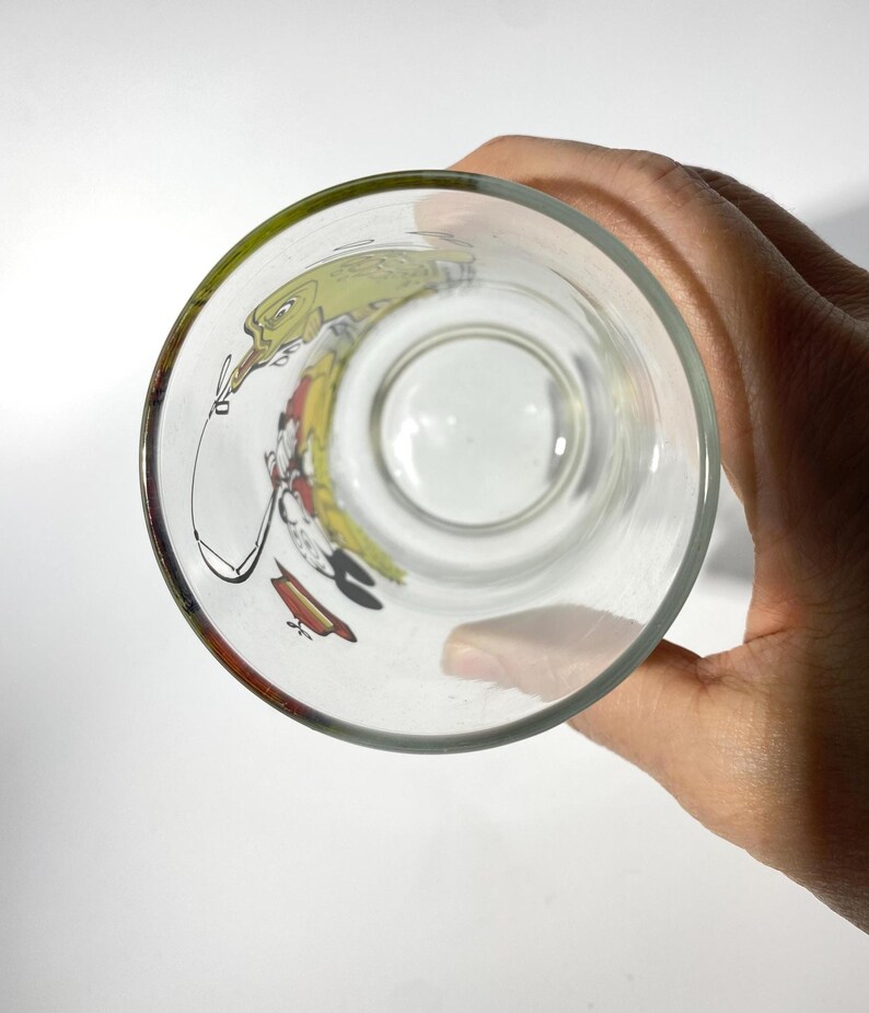 K&ouml;nnte beinhalten: Klares Glas mit einem Cartoon-Charakter-Design. Das Glas hat einen gr&uuml;nen Rand und eine farbenfrohe Illustration auf der Innenseite. Das Glas wird von einer Hand gehalten.
