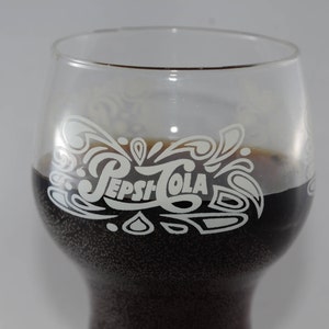 May include: A glass with a white Pepsi-Cola logo etched on the side, filled with a dark brown liquid. The glass has a rounded bottom and a wide mouth.