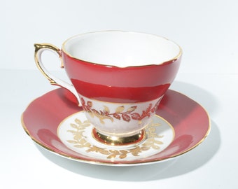 Può includere: Una tazza da tè rossa e un piattino con un motivo a foglie dorate. La tazza e il piattino sono entrambi rifiniti in oro.
