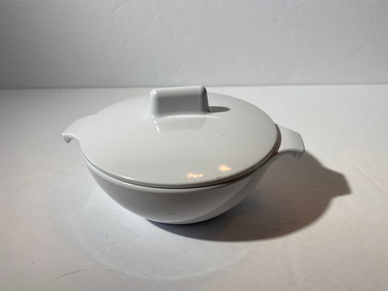 May include: White ceramic bowl with a lid. The bowl has a rounded shape and a small handle on the lid.