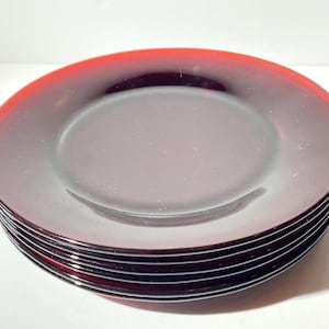 Puede incluir: Una pila de seis platos redondos con un esquema de color degradado. Los platos pasan de un rojo intenso en los bordes a un centro más claro y translúcido. Los platos son de vidrio y están apilados ordenadamente.