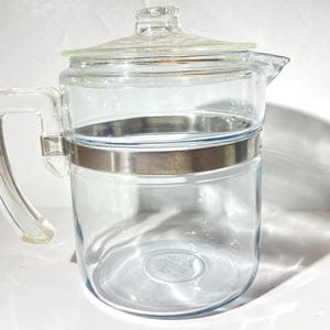 Może przedstawiać: Przezroczysty szklany dzbanek do kawy z pokrywką i uchwytem. Dzbanek ma srebrzysty metalowy pasek pośrodku. Uchwyt jest przezroczysty i przymocowany do boku. Pokrywka jest również przezroczysta.