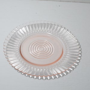 Puede incluir: Un plato de vidrio transparente con un centro rosa y un borde decorativo y acanalado. El centro del plato presenta c&iacute;rculos conc&eacute;ntricos. El plato es probablemente para servir alimentos.
